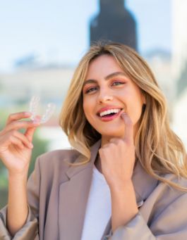 Blonde woman holding a clear aligner and pointing to her smile